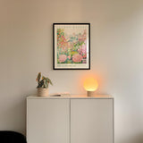 a white cabinet with a potted plant and a lamp on top, set against a wall with a framed poster of a garden scene hanging above it.