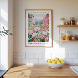 a kitchen scene with a framed poster of a flower market in Greenwich, England, hanging on the wall above a wooden counter. The poster displays a colorful illustration of flowers and plants, and the text "FLOWER MARKET GREENWICH" is visible. On the counter, there is a bowl of lemons, and the wall has white tiles.