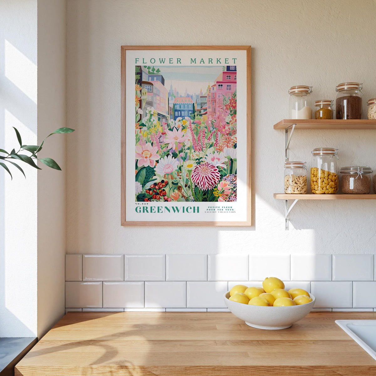 a kitchen scene with a framed poster of a flower market in Greenwich, England, hanging on the wall above a wooden counter. The poster displays a colorful illustration of flowers and plants, and the text "FLOWER MARKET GREENWICH" is visible. On the counter, there is a bowl of lemons, and the wall has white tiles.