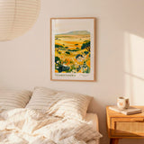 a bedroom scene with a framed print of a landscape depicting a field of flowers hanging on the wall above a bed with striped pillows.