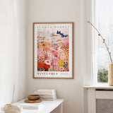 a framed poster of a colorful floral scene hanging on a wall, with a white table and a vase of flowers nearby.