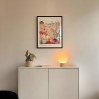 a white cabinet with a potted plant and a lamp on top, set against a wall with a framed poster of a cityscape above it.