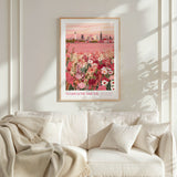 a framed poster of a cityscape with a pink and green floral background, hanging on a white wall above a white couch with a blanket draped over it.