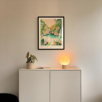 a white cabinet with a plant and a lamp on top, and a framed picture of a tropical landscape hanging on the wall above it.