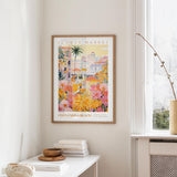 a framed poster of a colorful, tropical-themed flower market, hanging on a wall in a room with a window and a small table with books and a vase.