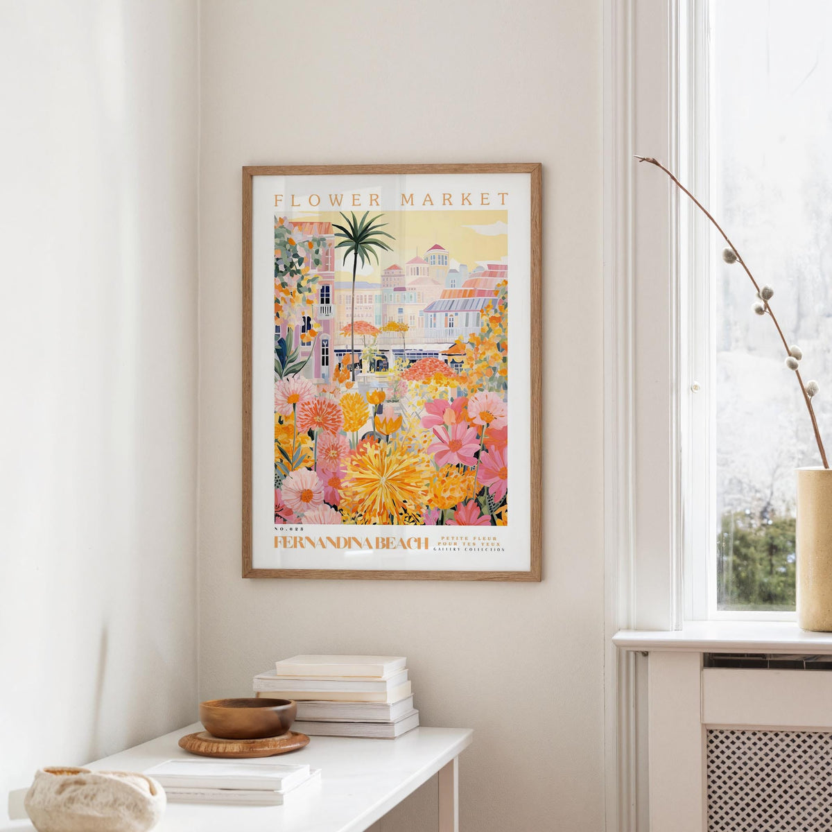 a framed poster of a colorful, tropical-themed flower market, hanging on a wall in a room with a window and a small table with books and a vase.
