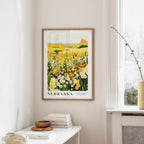 a framed poster of a field of flowers hanging on a wall, with a window and a small table with books and a vase nearby.