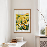 a framed poster of a field of flowers hanging on a wall, with a window and a small table with books and a vase nearby.