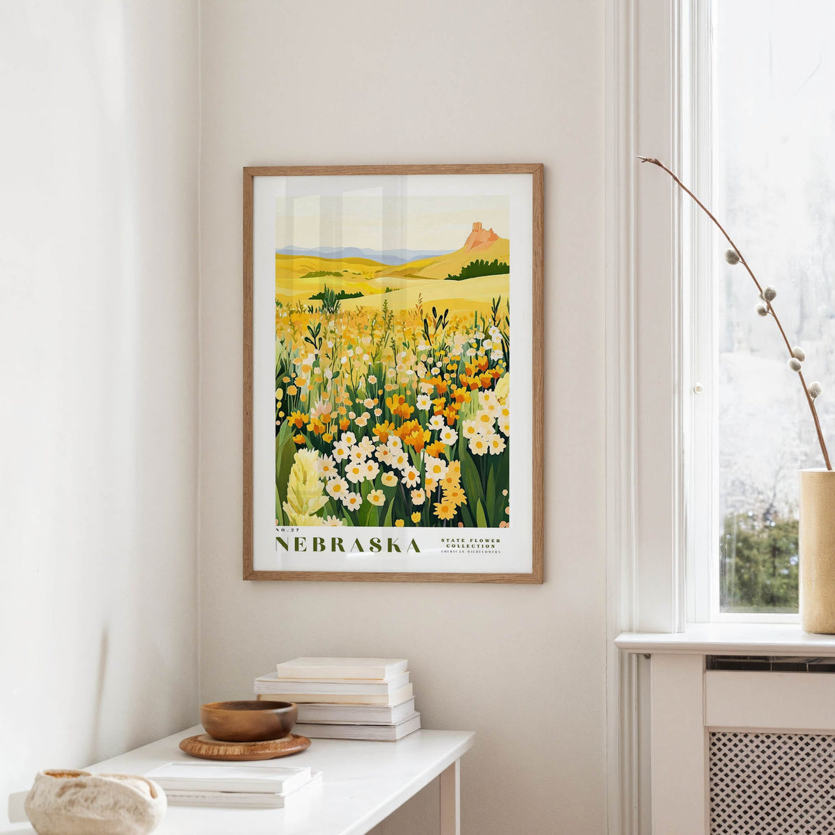 a framed poster of a field of flowers hanging on a wall, with a window and a small table with books and a vase nearby.
