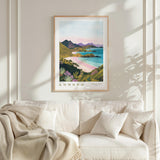 a framed poster of a scenic landscape with mountains, a beach, and a body of water, hanging on a wall above a white couch with pillows.