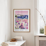 a framed poster of a coastal scene with a house, boats, and flowers, hanging on a wall in a room with a window and a table with books and a vase.