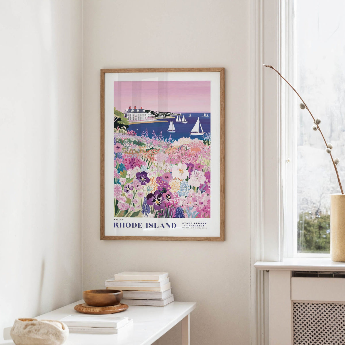 a framed poster of a coastal scene with a house, boats, and flowers, hanging on a wall in a room with a window and a table with books and a vase.