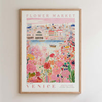 The image is a framed poster depicting a vibrant and colorful scene from Venice, Italy. The poster showcases the city's iconic architecture, including the Ponte Vecchio, surrounded by a lush garden filled with flowers and greenery. The poster is framed with a wooden border and features the text "FLOWER MARKET" and "VENICE" prominently displayed.