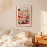 a bedroom scene with a framed poster of a colorful flower market in Kansas City hanging on the wall above a bed with striped pillows.