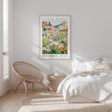 a bedroom with a bed, a woven chair, and a framed poster of a flower market in Monterey.