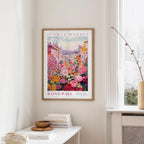 a framed poster of a colorful floral scene hanging on a wall, with a window and a small table with books and a vase nearby.