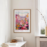 a framed poster of a colorful floral scene hanging on a wall, with a window and a small table with books and a vase nearby.