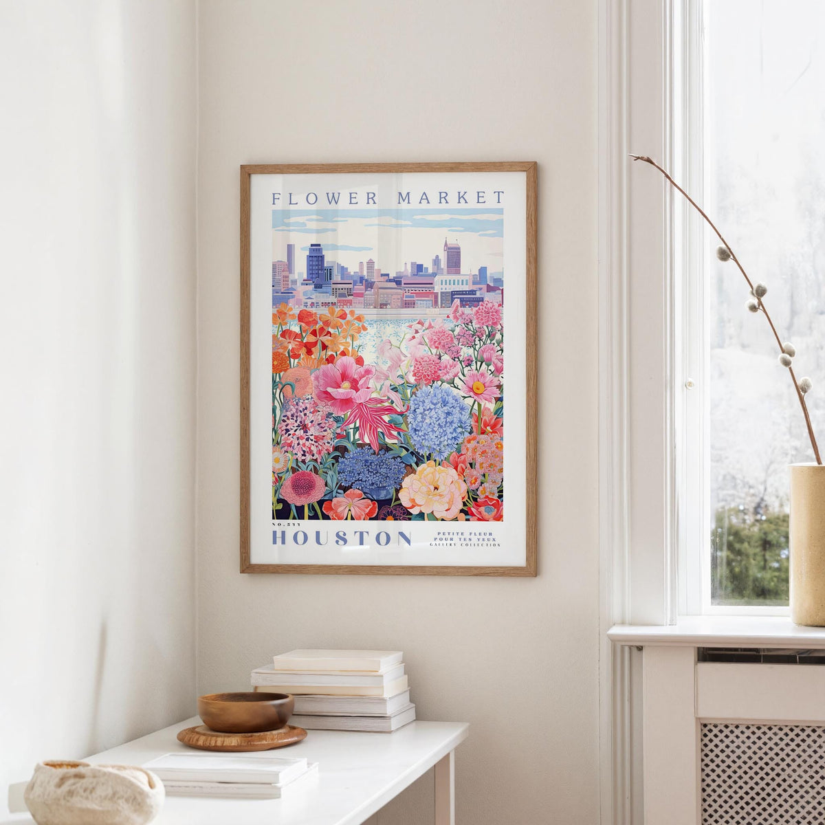 a framed poster of a colorful floral scene hanging on a wall, with a window and a small table with books and a vase in the foreground.