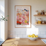 a framed poster of a colorful floral scene hanging on a wall above a kitchen counter with a bowl of lemons on it.