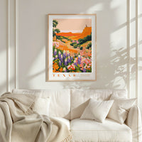 a framed poster of a colorful landscape with flowers hanging on a wall above a white couch with pillows.