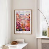 a framed poster of a colorful floral scene hanging on a wall, with a white table and a vase of flowers in the foreground.