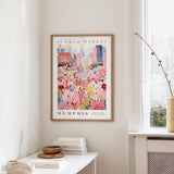 a framed poster of a colorful cityscape with a flower market scene, hanging on a wall in a room with a window and a small table with books and a bowl.