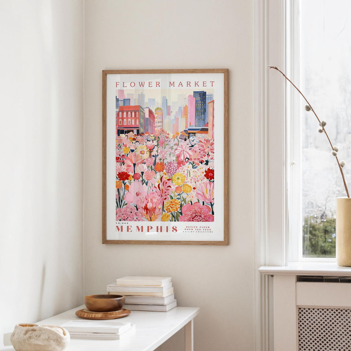 a framed poster of a colorful cityscape with a flower market scene, hanging on a wall in a room with a window and a small table with books and a bowl.