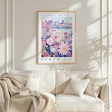 a framed poster of a cityscape with a flower market in the background, hanging on a wall above a white couch with a blanket draped over it.