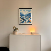 a framed poster of a mountain landscape, a potted plant, and a small lamp on a white cabinet.