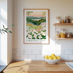 a framed poster of a garden with white flowers hanging on a wall above a wooden table with a bowl of lemons.