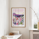 a framed poster of a colorful floral landscape hanging on a wall, with a white table and a vase of flowers in the foreground.
