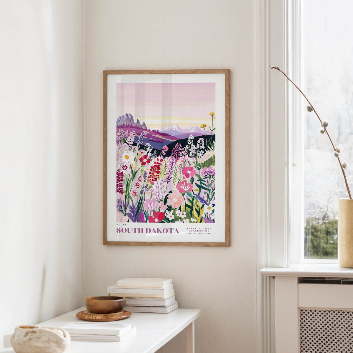 a framed poster of a colorful floral landscape hanging on a wall, with a white table and a vase of flowers in the foreground.