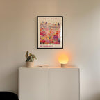 a white cabinet with a potted plant and a lamp on top, and a framed poster of a flower market hanging on the wall above it.