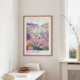 a framed poster of a colorful garden scene hanging on a wall, with a white table and a vase of flowers in the foreground.