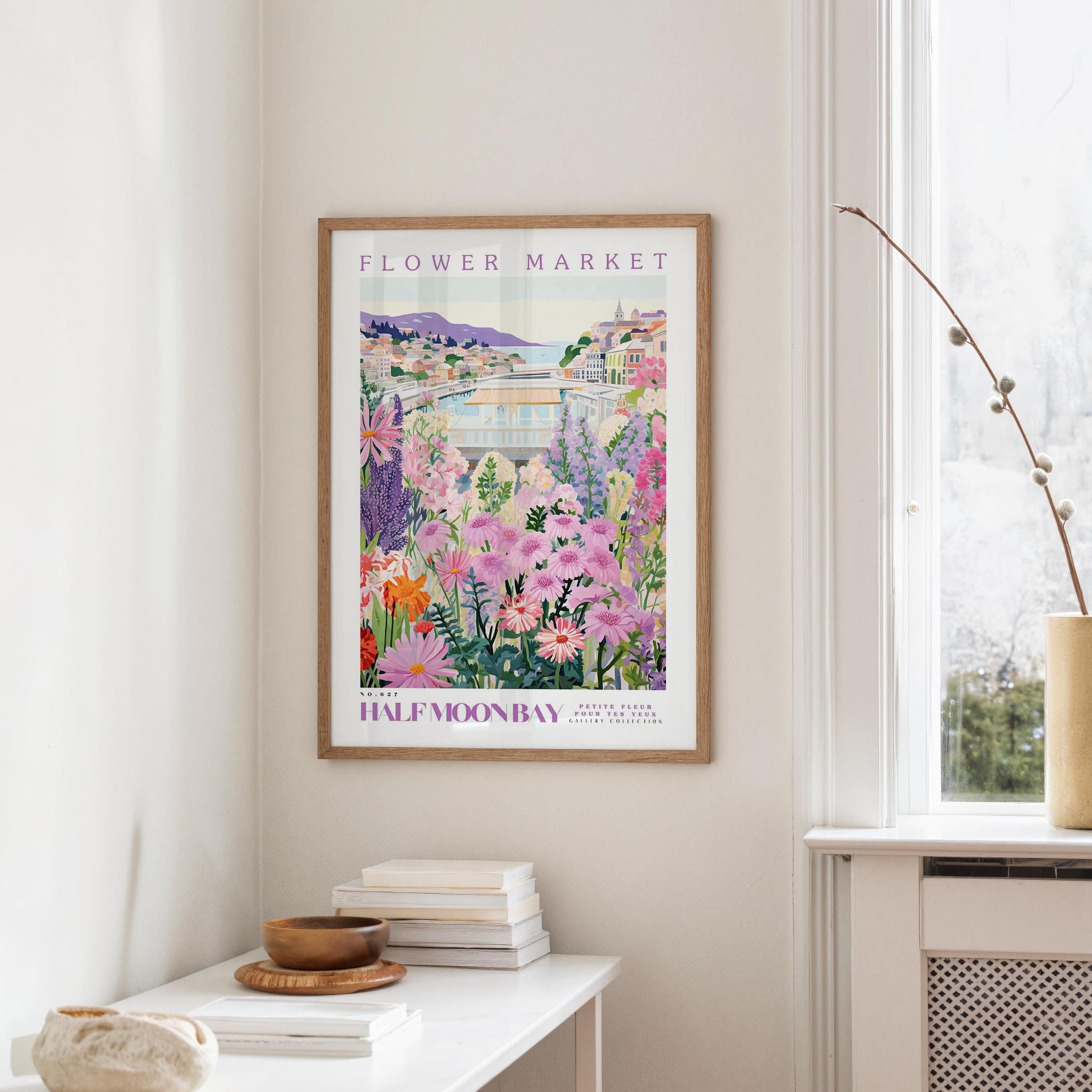 a framed poster of a colorful garden scene hanging on a wall, with a white table and a vase of flowers in the foreground.