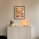 a white cabinet with a plant and a lamp on top, and a framed poster of a desert landscape hanging on the wall above it.