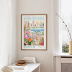 a framed poster of a colorful floral scene hanging on a wall, with a window and a small table with books and a vase in the foreground.