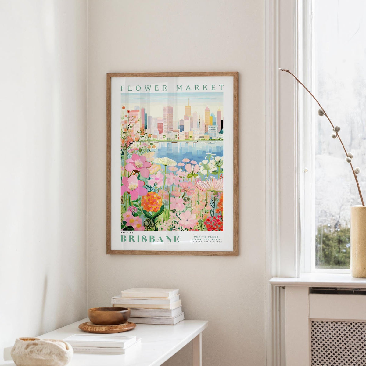 a framed poster of a colorful floral scene hanging on a wall, with a window and a small table with books and a vase in the foreground.