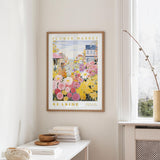 a framed poster of a colorful flower market scene hanging on a wall, with a white table and a vase of flowers nearby.