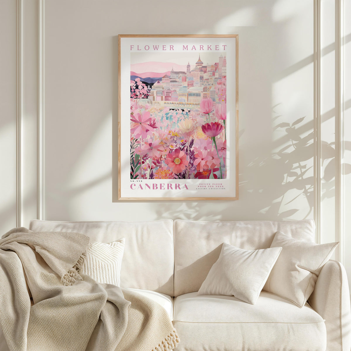 a framed poster of a cityscape with a pink and white color scheme, hanging on a wall above a white couch with a blanket draped over it.