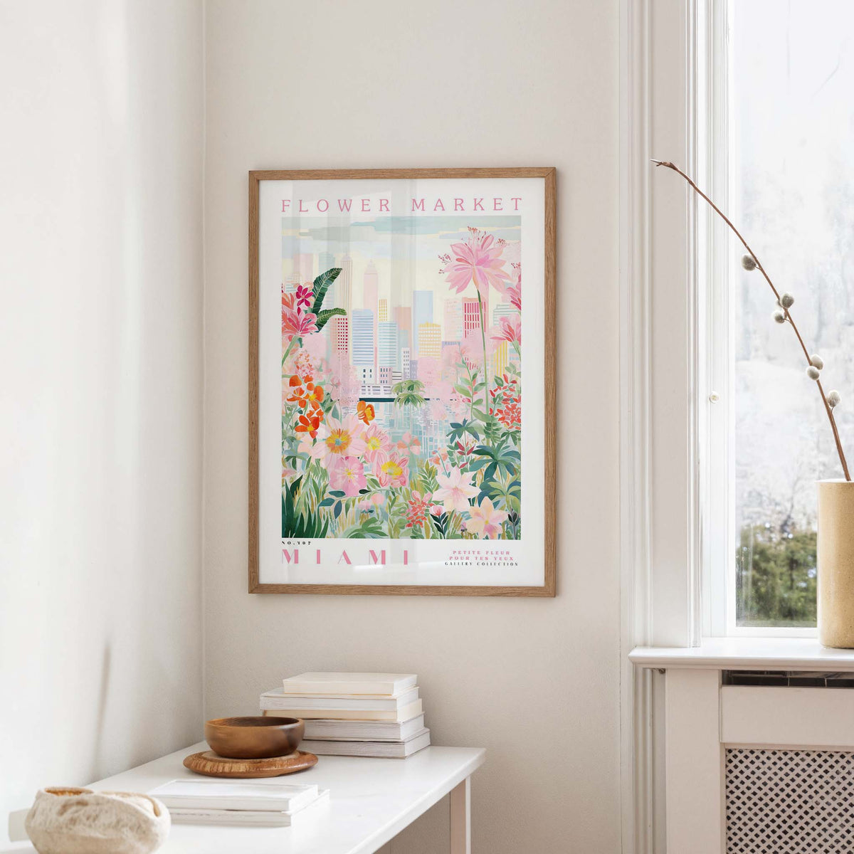 a framed poster of a colorful floral scene hanging on a wall, with a white table and a vase of flowers in the foreground.
