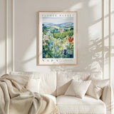 a framed poster of a field of flowers hanging on a wall above a white couch with a blanket draped over it.