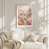 a poster of a cityscape with a flower market in Portland, Oregon, hanging on a wall above a white couch with a blanket draped over it.