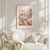 a poster of a cityscape with a flower market in Portland, Oregon, hanging on a wall above a white couch with a blanket draped over it.
