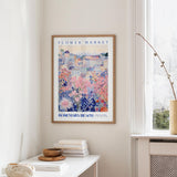 a framed poster of a colorful floral scene hanging on a wall, with a white table and a vase of flowers nearby.