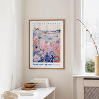 a framed poster of a colorful floral scene hanging on a wall, with a white table and a vase of flowers nearby.
