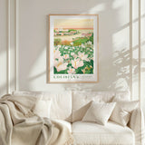 a framed poster of a field of flowers hanging on a wall above a white couch with pillows.