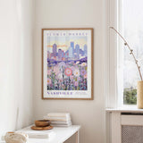 a framed poster of a cityscape with a field of flowers in the foreground, hanging on a wall in a room with a window and a table with books and a vase.