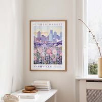 a framed poster of a cityscape with a field of flowers in the foreground, hanging on a wall in a room with a window and a table with books and a vase.