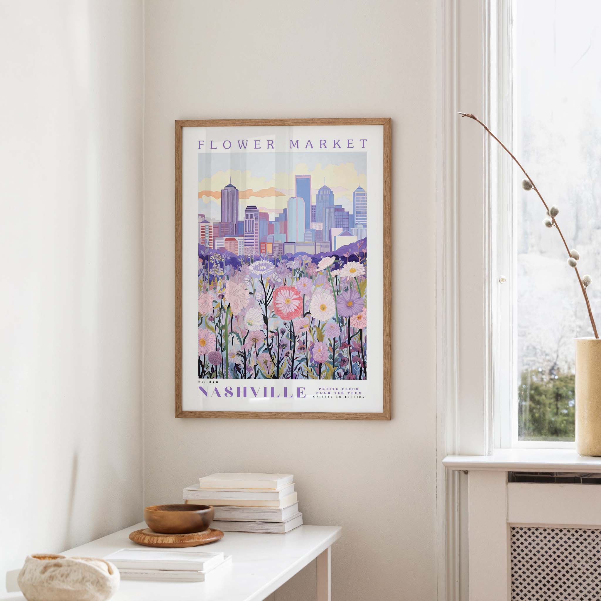 a framed poster of a cityscape with a field of flowers in the foreground, hanging on a wall in a room with a window and a table with books and a vase.